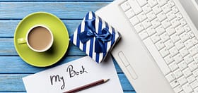 image of laptop, cup of coffee, pen, open journal titled my book, and gift wrapped in white and blue paper with a blue ribbon showing different options for gifts for writers