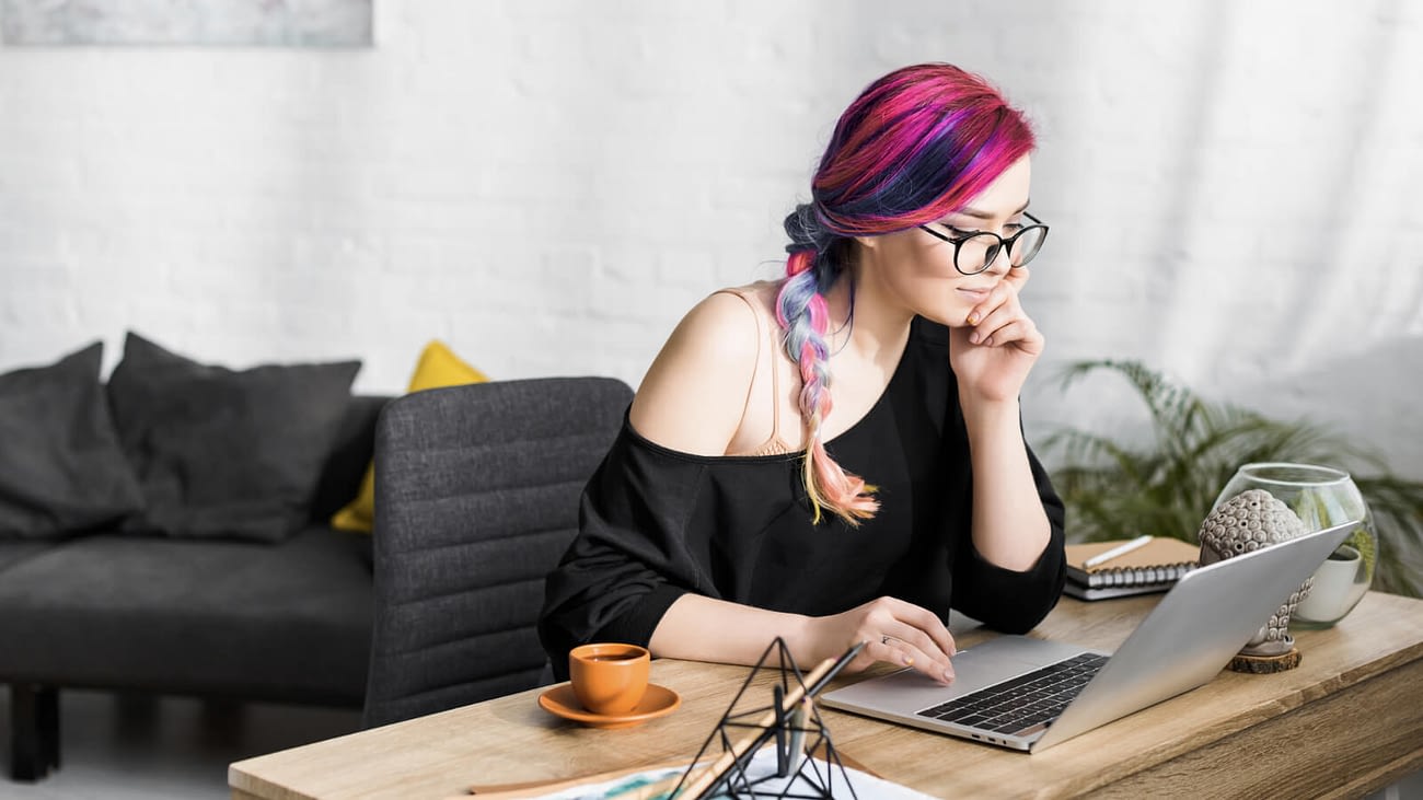 femme person with pink and purple hair sitting at table looking laptop while writing
