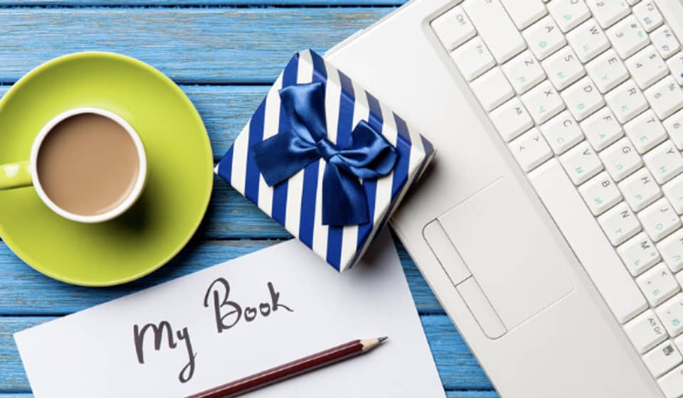 image of laptop, cup of coffee, pen, open journal titled my book, and gift wrapped in white and blue paper with a blue ribbon showing different options for gifts for writers