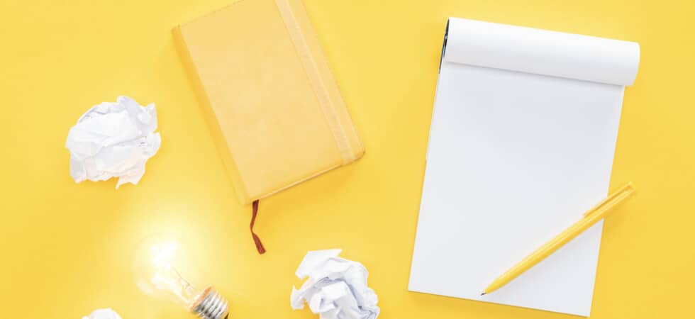 concept of writing ideas on yellow desk with yellow notebook, pad of paper, yellow pen, crumpled papers, and a lit up lightbulb